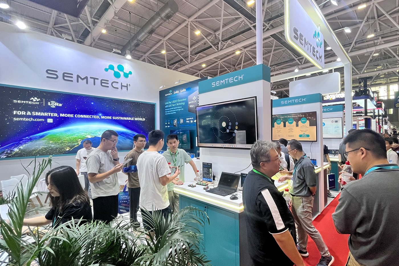 Semtech exhibition booth at IOTE Shenzhen 2025 trade show with multiple attendees gathered around interactive demonstration displays, featuring the company's logo, large digital screens showing product demos, and booth staff engaging with visitors in a modern white and teal-colored booth setup