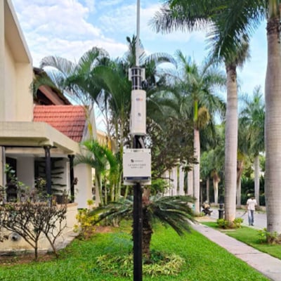 LoRa-BLOG-Smart Meters India-Part LoRaWAN gateway installation on a pole in Palm Meadows residential community, featuring the communication device mounted on a black pole surrounded by palm trees and landscaped grounds with houses visible in the background. This IoT infrastructure enables Enthu Technology's smart energy and water management system that provides real-time monitoring capabilities and improved resource efficiency for the residential community.