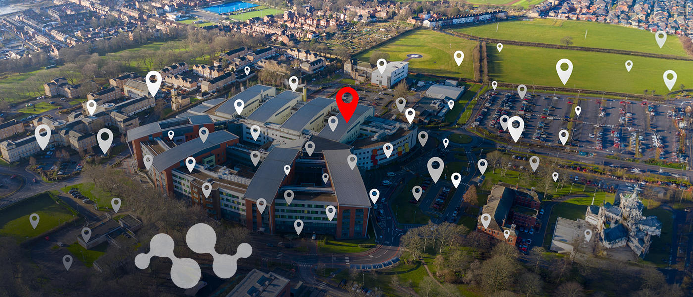 Aerial view of a modern building complex with numerous white location pin markers scattered across the campus and one prominent red location pin marker, illustrating LoRa-based asset tracking and location intelligence technology in an urban development setting