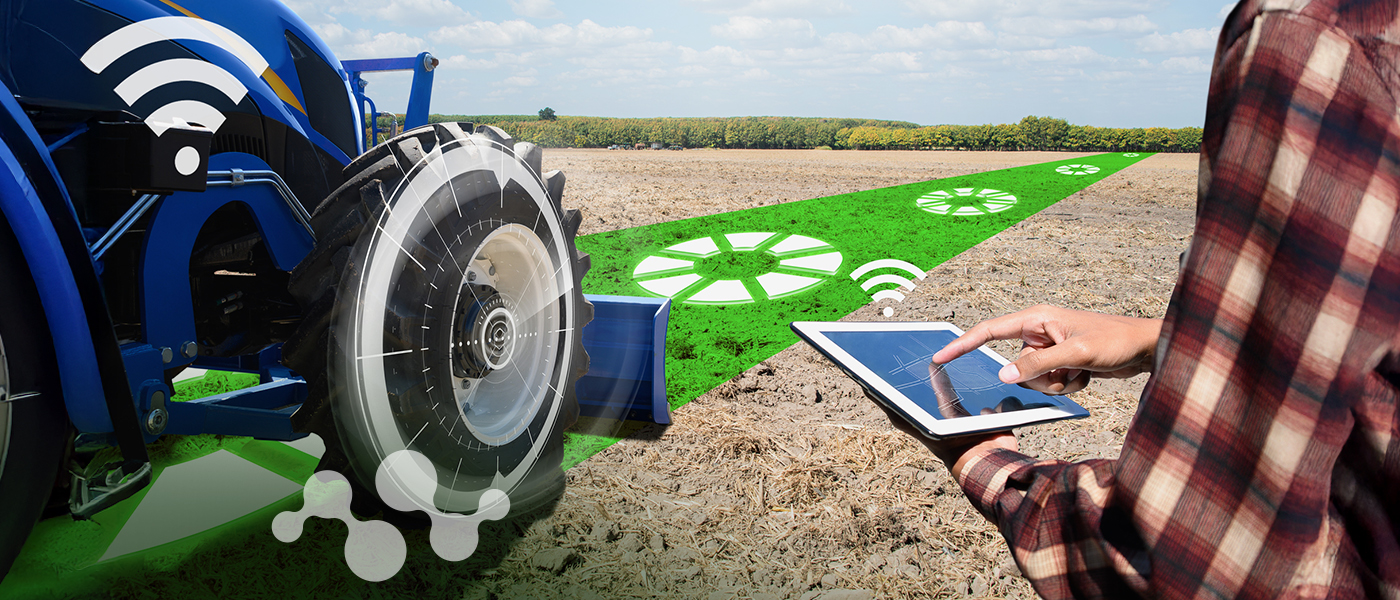 A farmer in a plaid shirt operates a tablet device to control a blue tractor in a prepared agricultural field. Digital overlay graphics show WiFi connectivity symbols and gear icons scattered across the farmland, illustrating IoT-enabled smart farming technology and precision agriculture connectivity solutions